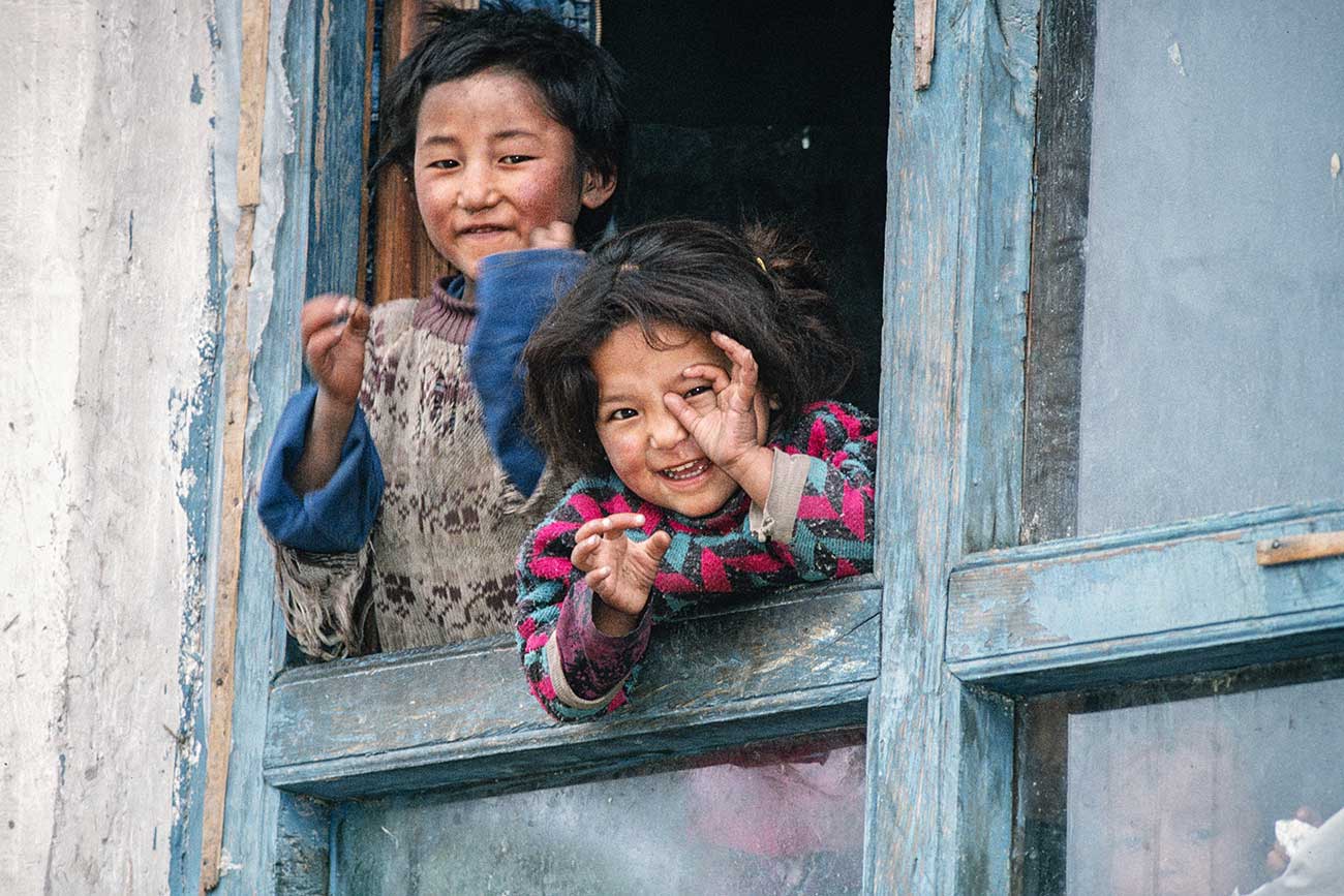 Deux enfants souriant à la fenêtre de leur maison de Padum, mimant le geste de faire des photos